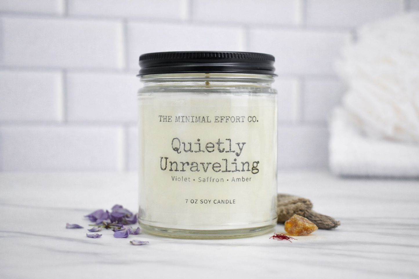 Candle jar labeled 'Quietly Unraveling' by The Minimal Effort Co. on a white surface with decorative elements.