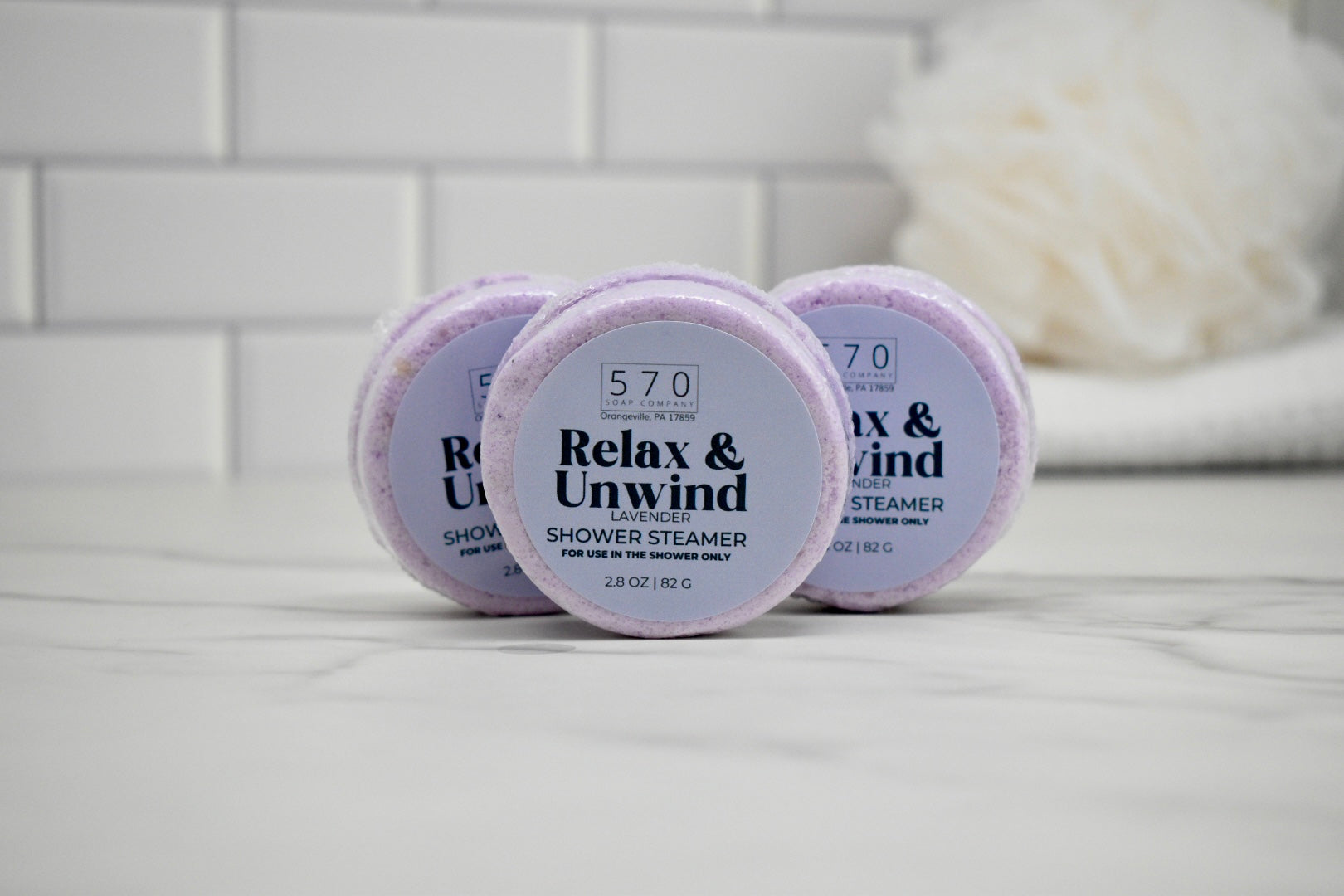 Three 'Relax & Unwind' shower steamer containers on a white surface with a blurred towel in the background.