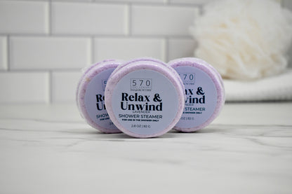 Three 'Relax & Unwind' shower steamer containers on a white surface with a blurred towel in the background.