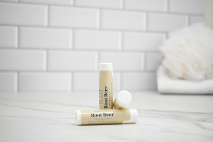 Two lip balm sticks labeled 'Root Beer' on a white surface with a tiled wall background.