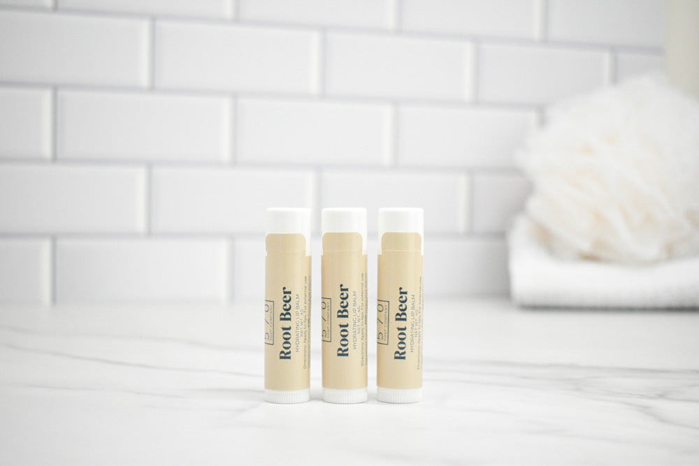 Three lip balm tubes labeled 'Root Beer' on a white surface with a tiled wall background.