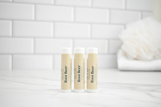 Three lip balm tubes labeled 'Root Beer' on a white surface with a tiled wall background.