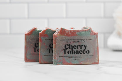 A trio of Cherry Tobacco Bar Soaps from 570 Soap Co. rests elegantly on a white marble surface. Their rustic packaging, showcasing text about the soap's crafting location, beautifully contrasts with the light tiled background. The inclusion of kaolin clay adds to its artisanal allure.