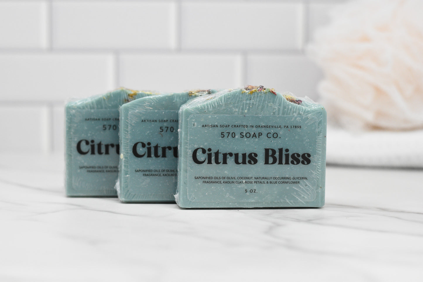 Three Citrus Bliss Soap Bars by 570 Soap Co. are elegantly wrapped and showcased on a marble countertop. The setting includes a white tiled wall in the background, along with a blurred white loofah, while the enticing aroma of Vibrant Citrus Fusion assures an indulgent moisturizing experience with each use.