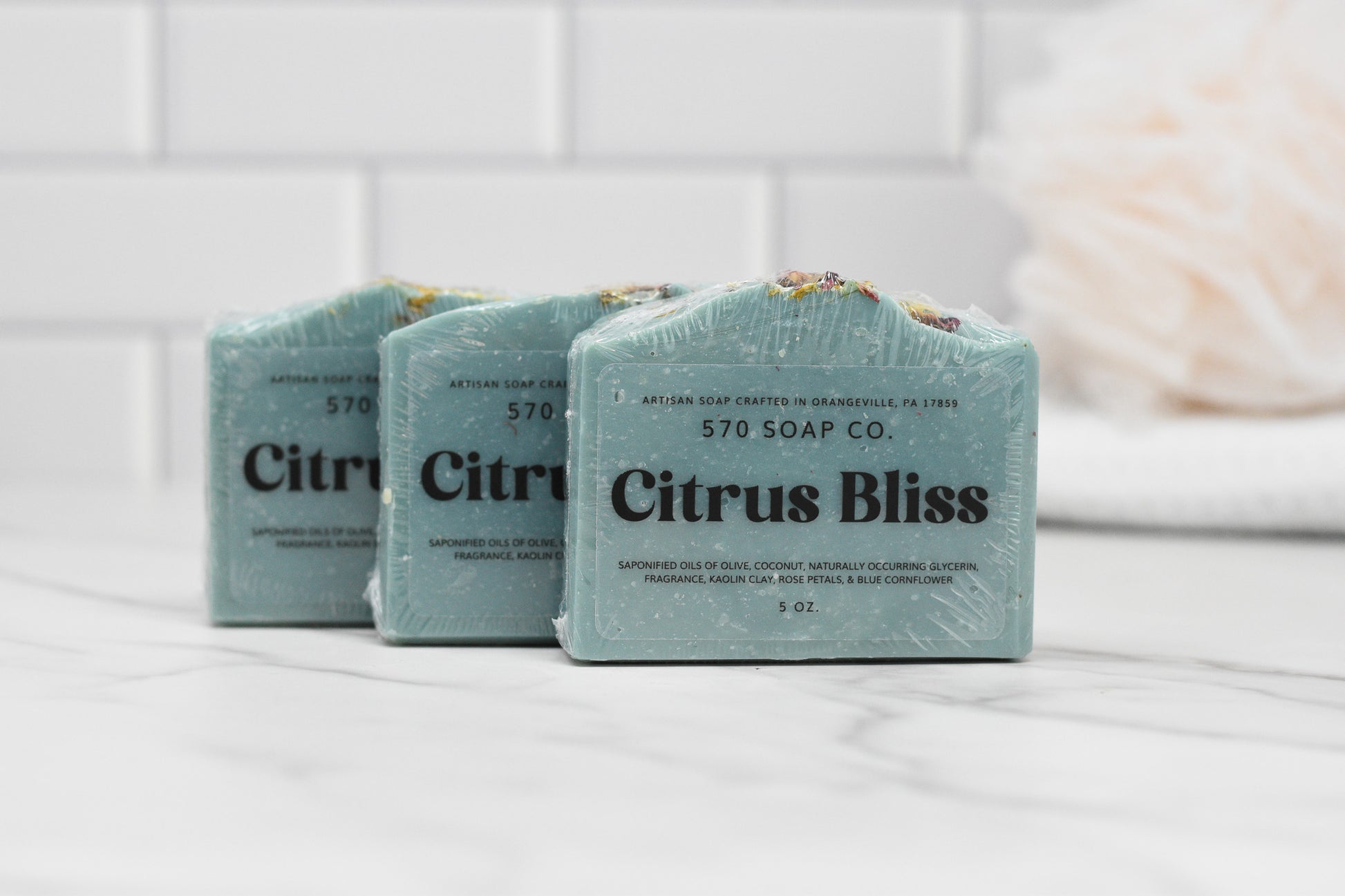 Three Citrus Bliss Soap Bars by 570 Soap Co. are elegantly wrapped and showcased on a marble countertop. The setting includes a white tiled wall in the background, along with a blurred white loofah, while the enticing aroma of Vibrant Citrus Fusion assures an indulgent moisturizing experience with each use.