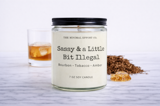 Candle labeled 'Sassy & a Little Bit Illegal' from The Minimal Effort Co. on a white surface with a glass of whiskey and tobacco leaves.