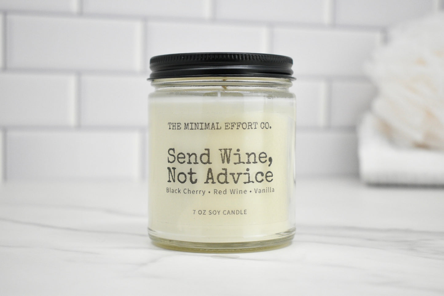 Candle jar labeled 'Send Wine, Not Advice' on a white surface with a tiled wall background.