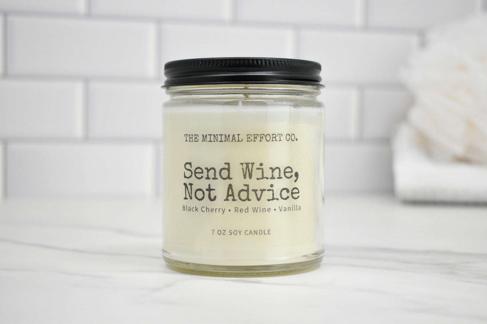 Candle jar labeled 'Send Wine, Not Advice' on a white surface with a tiled wall background.