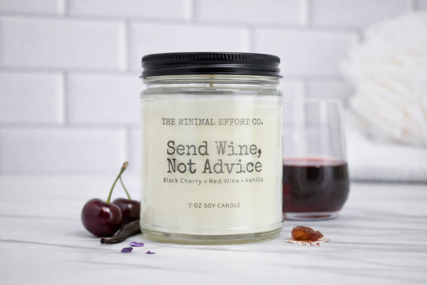 Candle labeled 'Send Wine, Not Advice' from The Minimal Effort Co. with a glass of red wine and cherries on a white surface.