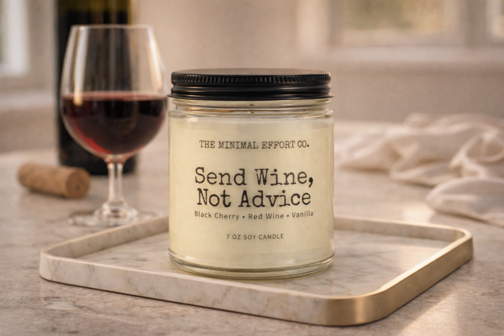 Candle labeled 'Send Wine, Not Advice' from The Minimal Effort Co. on a marble tray with a glass of red wine and bottle in the background.