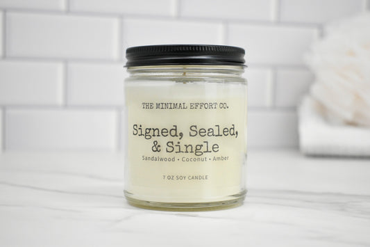 Candle jar labeled 'Signed, Sealed, & Single' by The Minimal Effort Co. on a white surface with a tiled wall background.