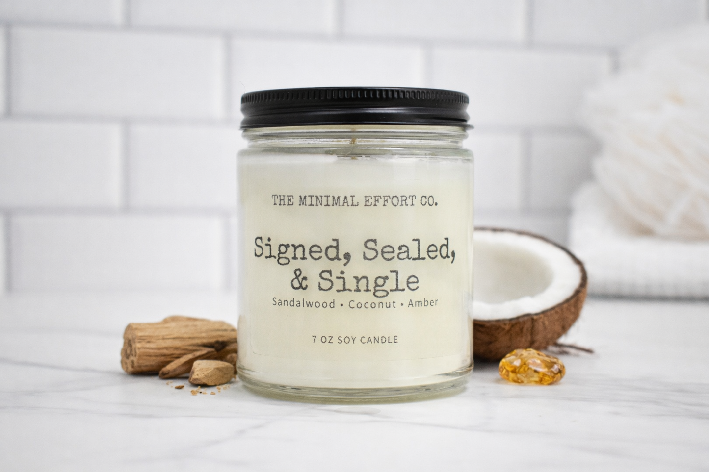 Candle labeled 'Signed, Sealed, & Single' from The Minimal Effort Co. on a white surface with a coconut and other items.