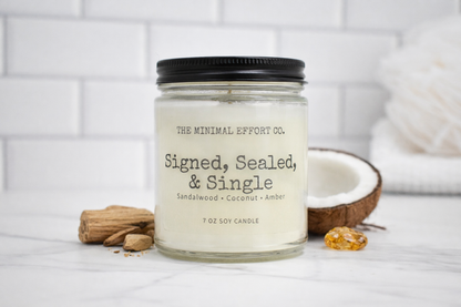 Candle labeled 'Signed, Sealed, & Single' from The Minimal Effort Co. on a white surface with a coconut and other items.