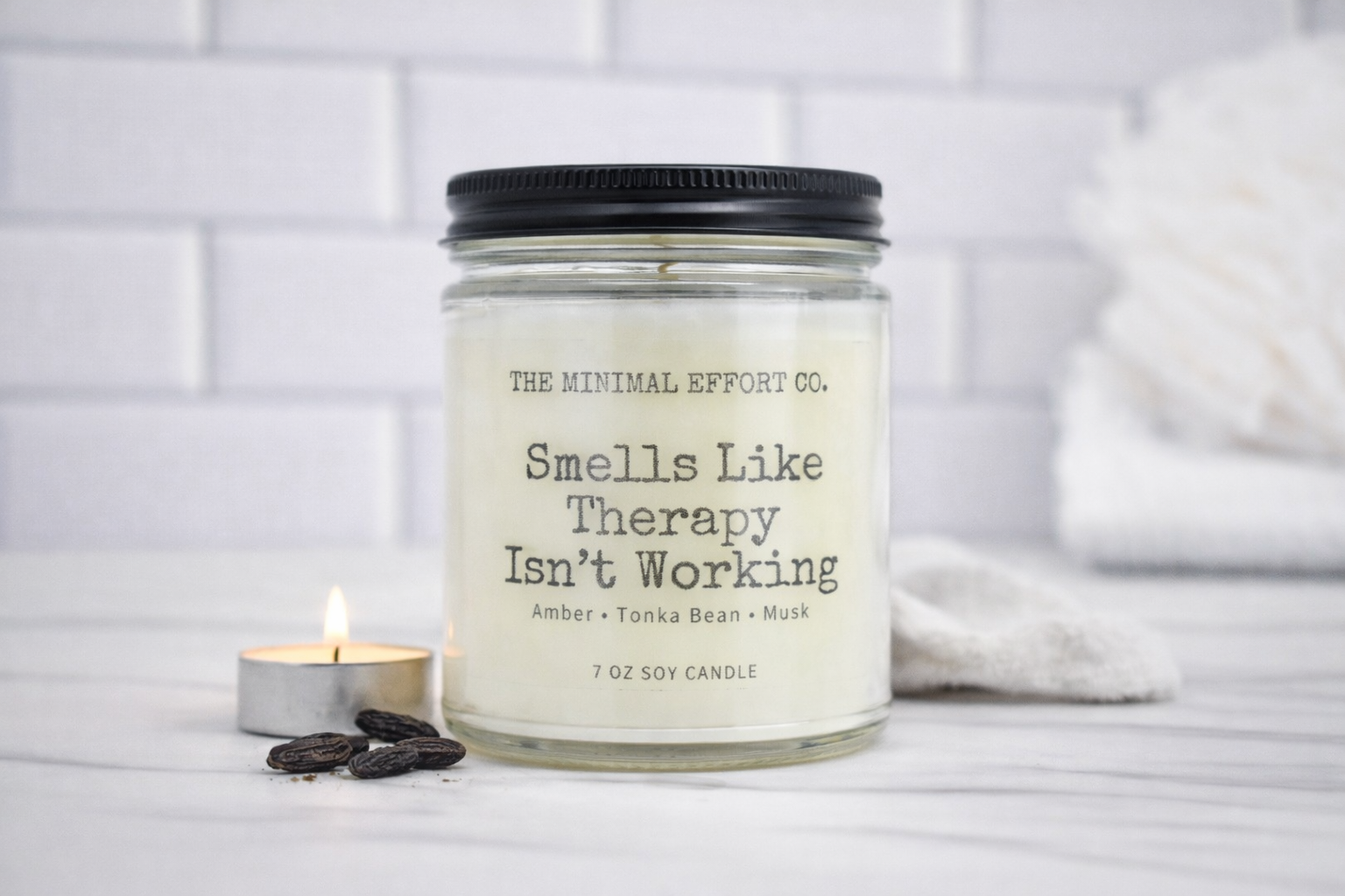 Candle jar labeled 'Smells Like Therapy Isn't Working' by The Minimal Effort Co. on a white surface with a blurred background.