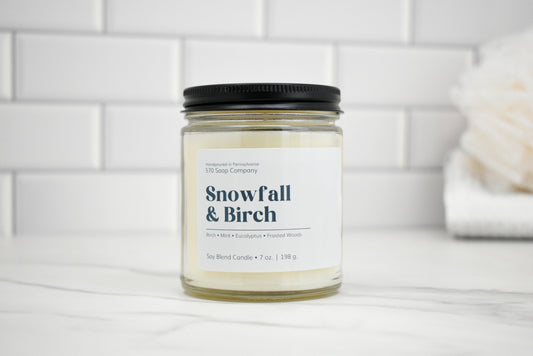 Candle jar labeled 'Snowfall & Birch' on a white surface with a tiled wall background