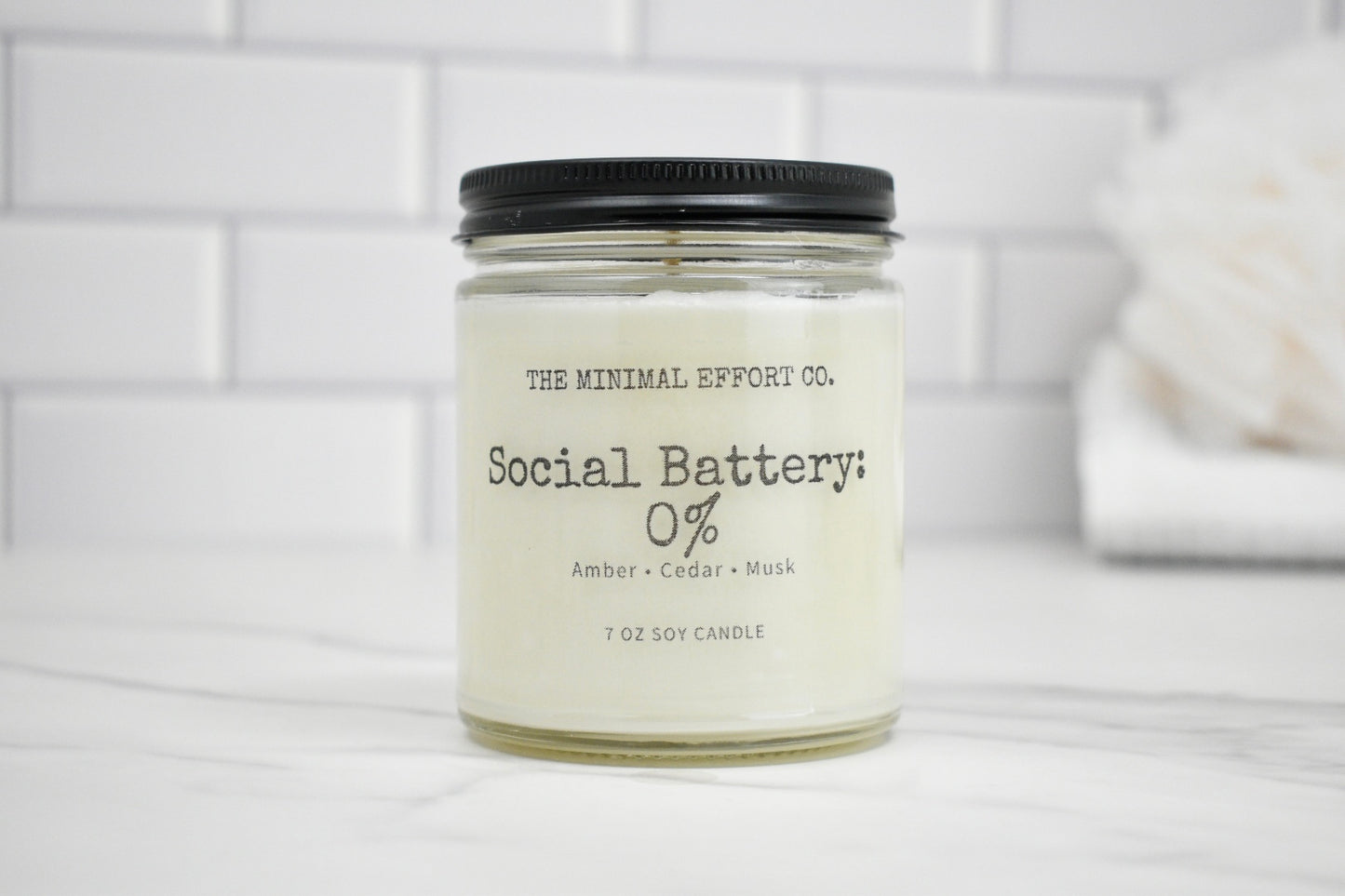 Candle jar labeled 'Social Battery 0%' by The Minimal Effort Co. on a white surface with a tiled wall background.