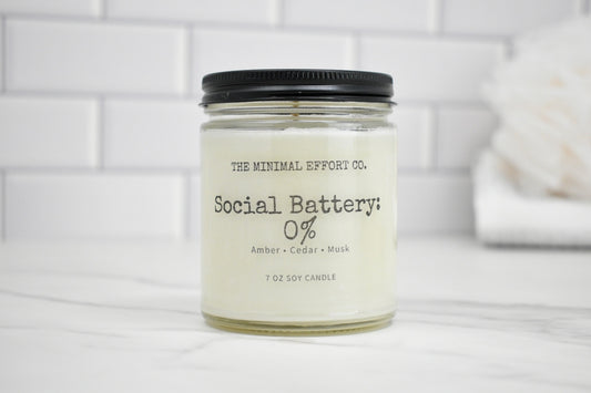 Candle jar labeled 'Social Battery 0%' by The Minimal Effort Co. on a white surface with a tiled wall background.