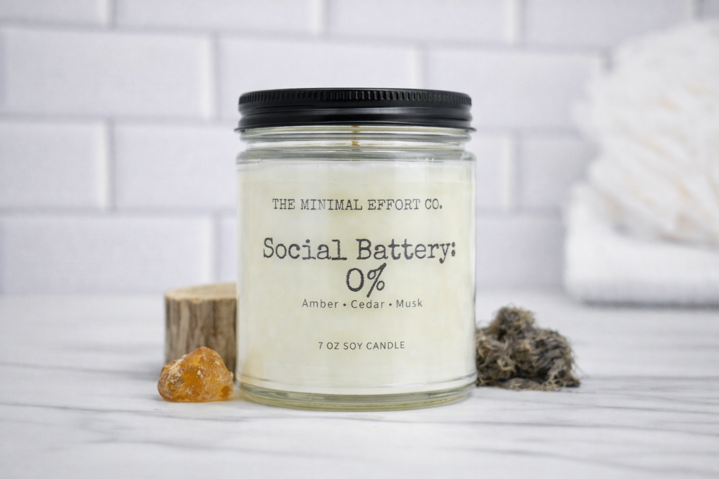 Candle labeled 'Social Battery: 0%' from 'The Minimal Effort Co.' on a white surface with a blurred background.