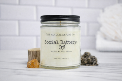 Candle labeled 'Social Battery: 0%' from 'The Minimal Effort Co.' on a white surface with a blurred background.