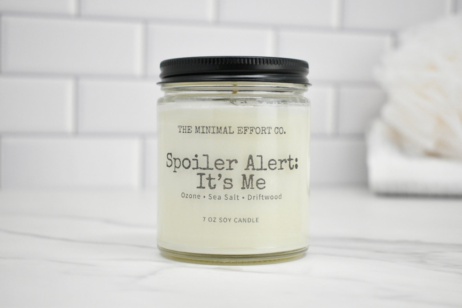 Candle jar labeled 'Spoiler Alert: It's Me' by The Minimal Effort Co. on a white surface with a tiled wall background.