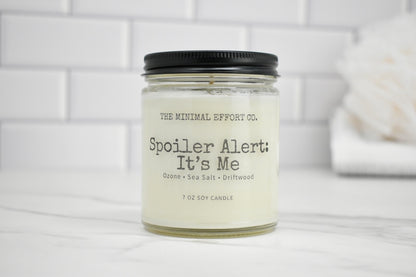 Candle jar labeled 'Spoiler Alert: It's Me' by The Minimal Effort Co. on a white surface with a tiled wall background.