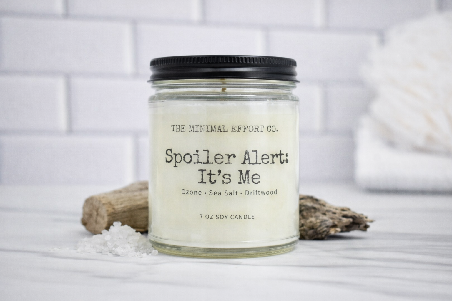 Candle with label 'Spoiler Alert: It's Me' from The Minimal Effort Co. on a white surface.
