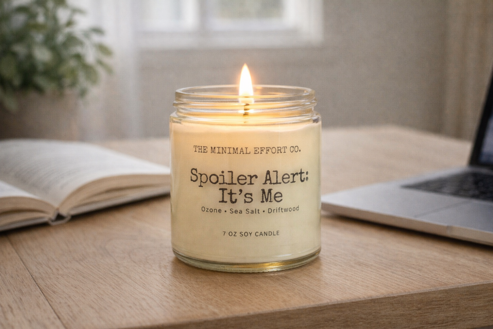 Candle labeled 'Spoiler Alert: It's Me' on a table with a laptop and book in the background