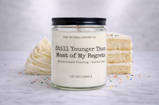 Candle labeled 'Still Younger Than Most of My Regrets' with a cake and frosting on a light background