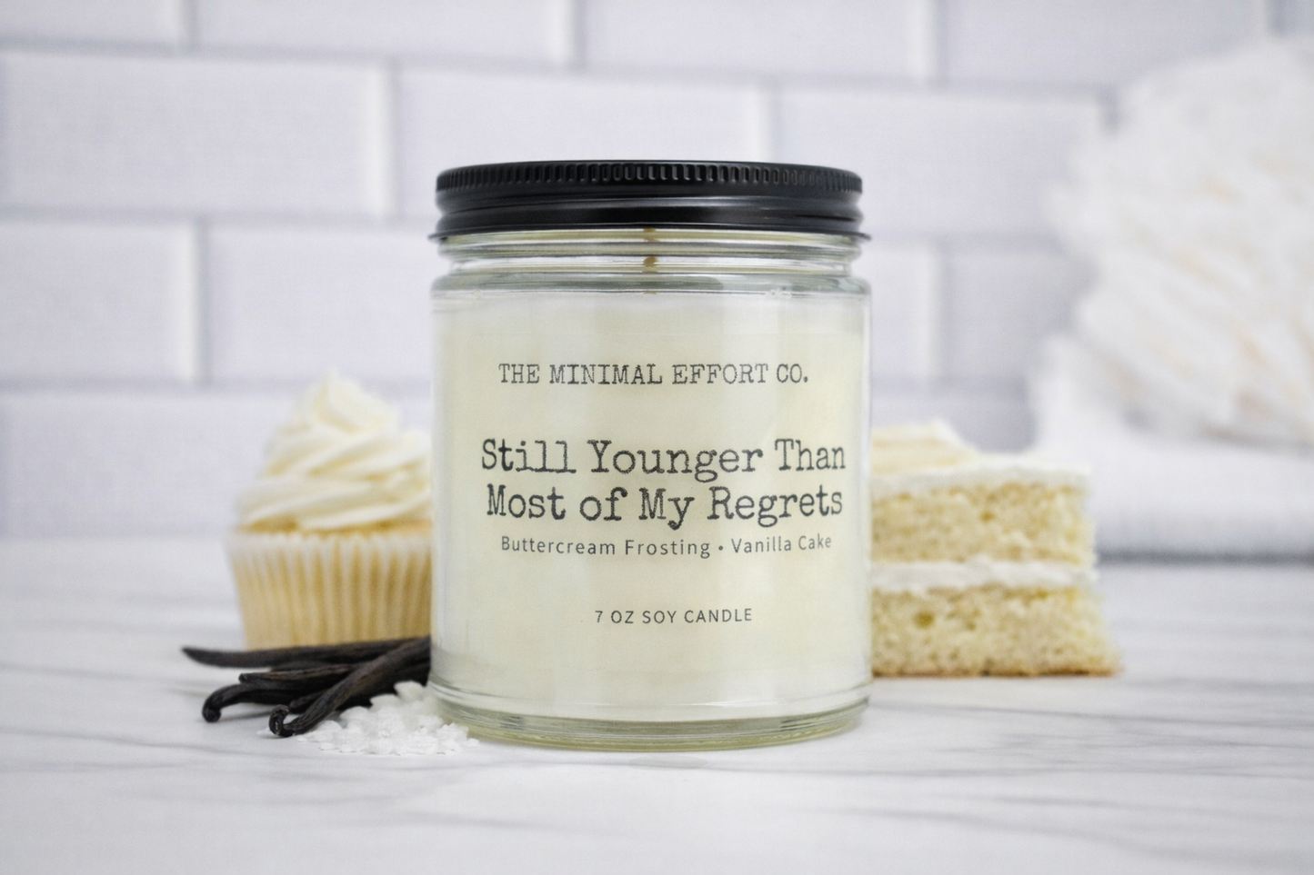 Candle with 'The Minimal Effort Co.' label on a white surface with vanilla beans and cupcakes.