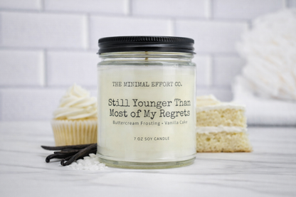 Candle with 'The Minimal Effort Co.' label on a white surface with vanilla beans and cupcakes.