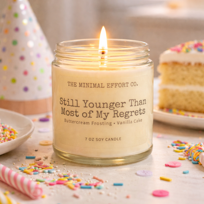 Candle in a glass jar with text, surrounded by birthday-themed items on a table.