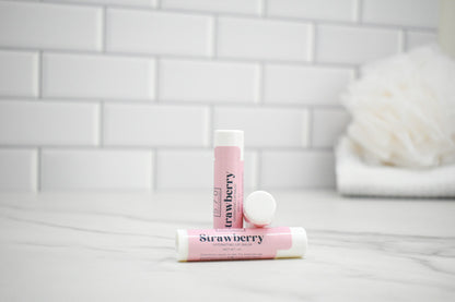 Two lip balm tubes labeled 'Strawberry' on a white surface with a tiled wall background.