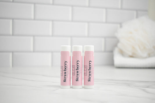 Three pink lip balms labeled 'Strawberry' on a white surface with a tiled wall background.