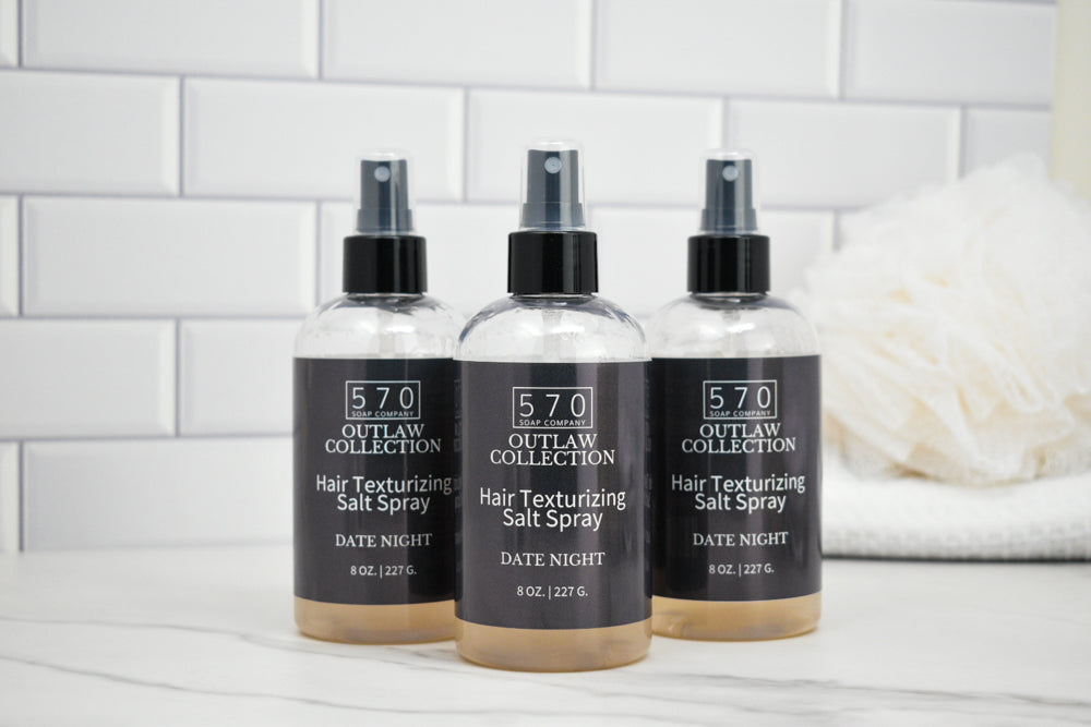 Three bottles of 570 Outlaw Collection hair texturizing salt spray on a white surface with a tiled wall background.
