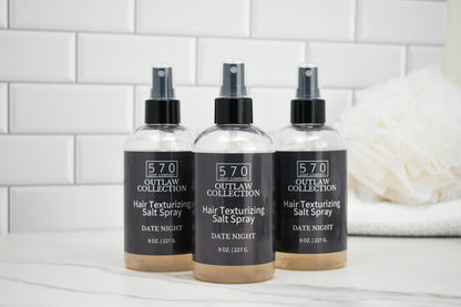 Three bottles of 570 Outlaw Collection hair texturizing salt spray on a white surface with a tiled wall background.