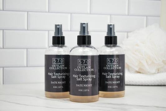Three bottles of 570 Outlaw Collection hair texturizing salt spray on a white surface with a tiled wall background.