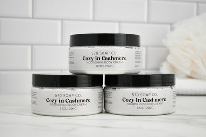 Three jars of Cozy in Cashmere body cream from 570 Soap Co. on a white surface with a tiled wall background.