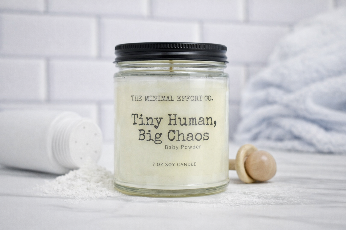 Candle with text 'Tiny Human, Big Chaos' by The Minimal Effort Co. on a white surface.