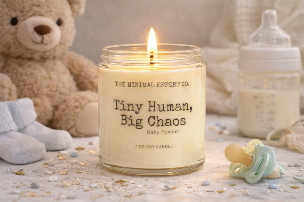 Candle with text 'Tiny Human, Big Chaos' by The Minimal Effort Co. in a setting with a teddy bear, baby bottle, and pacifier.