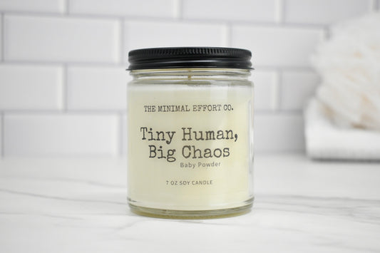 Candle jar labeled 'Tiny Human, Big Chaos' by The Minimal Effort Co. on a white surface with a tiled wall background.