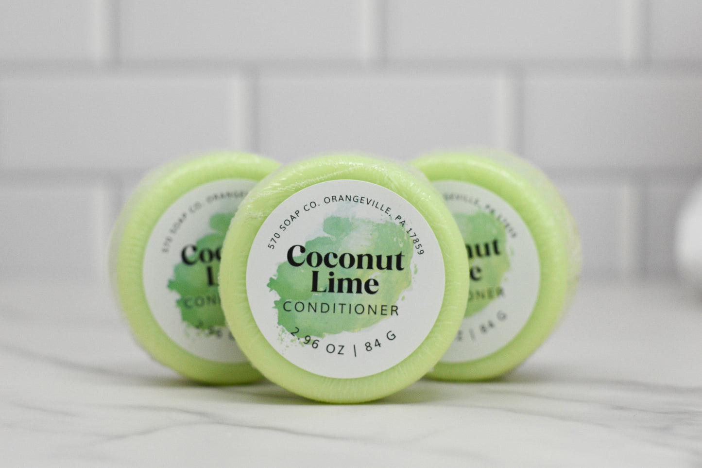 Three circular bars of the Coconut Lime Conditioner Bar for All Hair Types by 570 Soap Co. are stacked on a marble surface, offering Tropical Hydration. The packaging is light green, displaying the brand from Orangeville, PA. White subway tiles form the background, enhancing the refreshing allure.