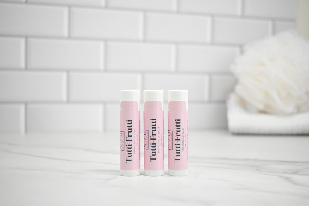Three Tootifrutti lip balm tubes on a white surface with a tiled wall background