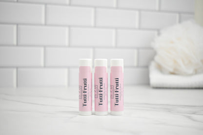 Three Tootifrutti lip balm tubes on a white surface with a tiled wall background