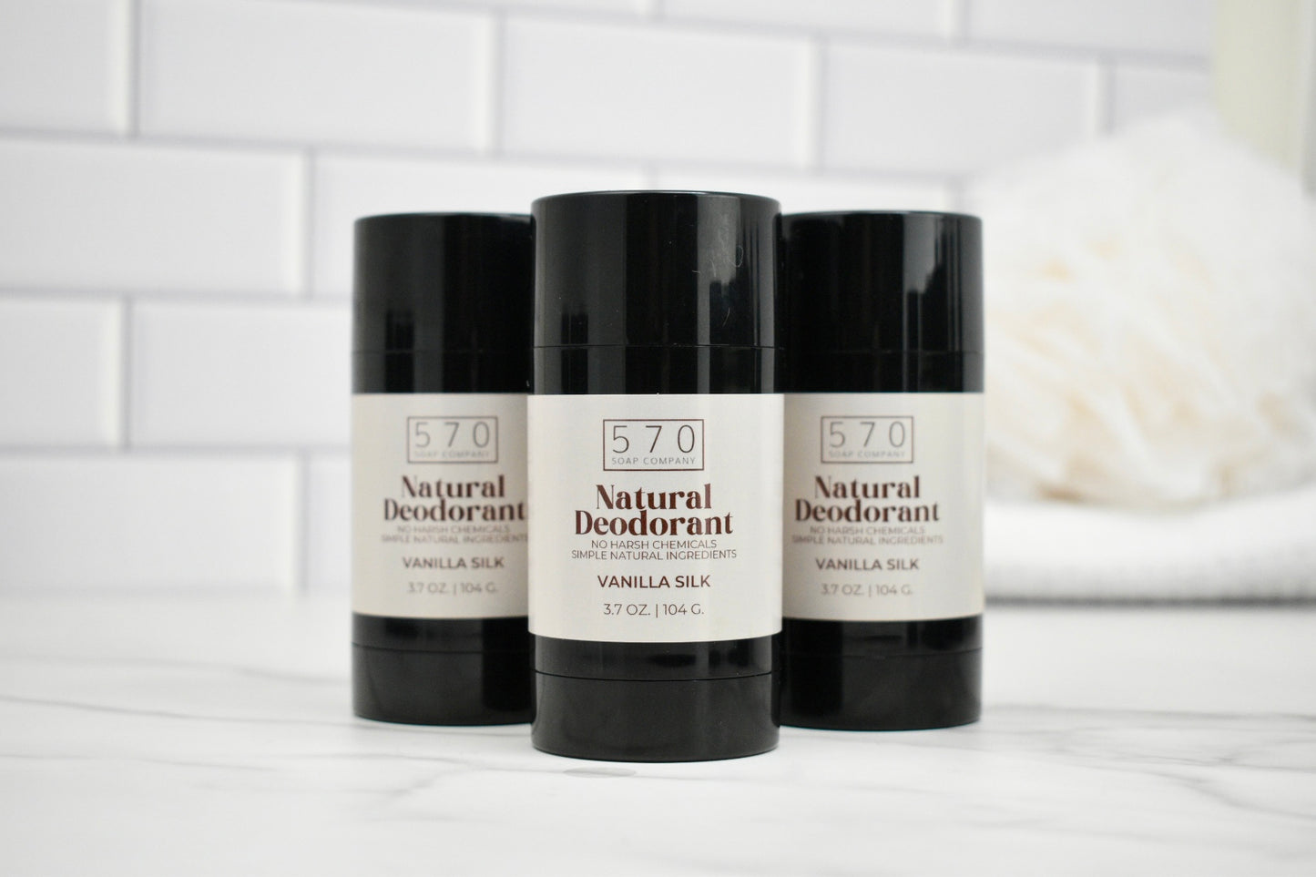 Three bottles of 570 Natural Deodorant on a white surface with a white tiled wall background.