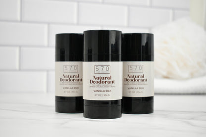 Three bottles of 570 Natural Deodorant on a white surface with a white tiled wall background.