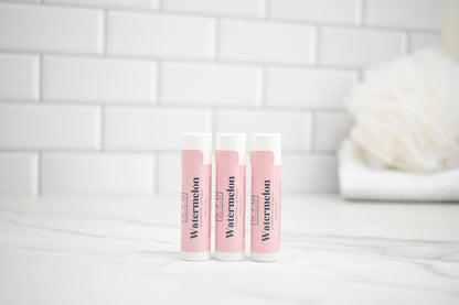 Three lip balm tubes labeled 'Watermelon' on a white surface with a tiled wall background.