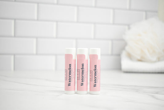 Three lip balm tubes labeled 'Watermelon' on a white surface with a tiled wall background.