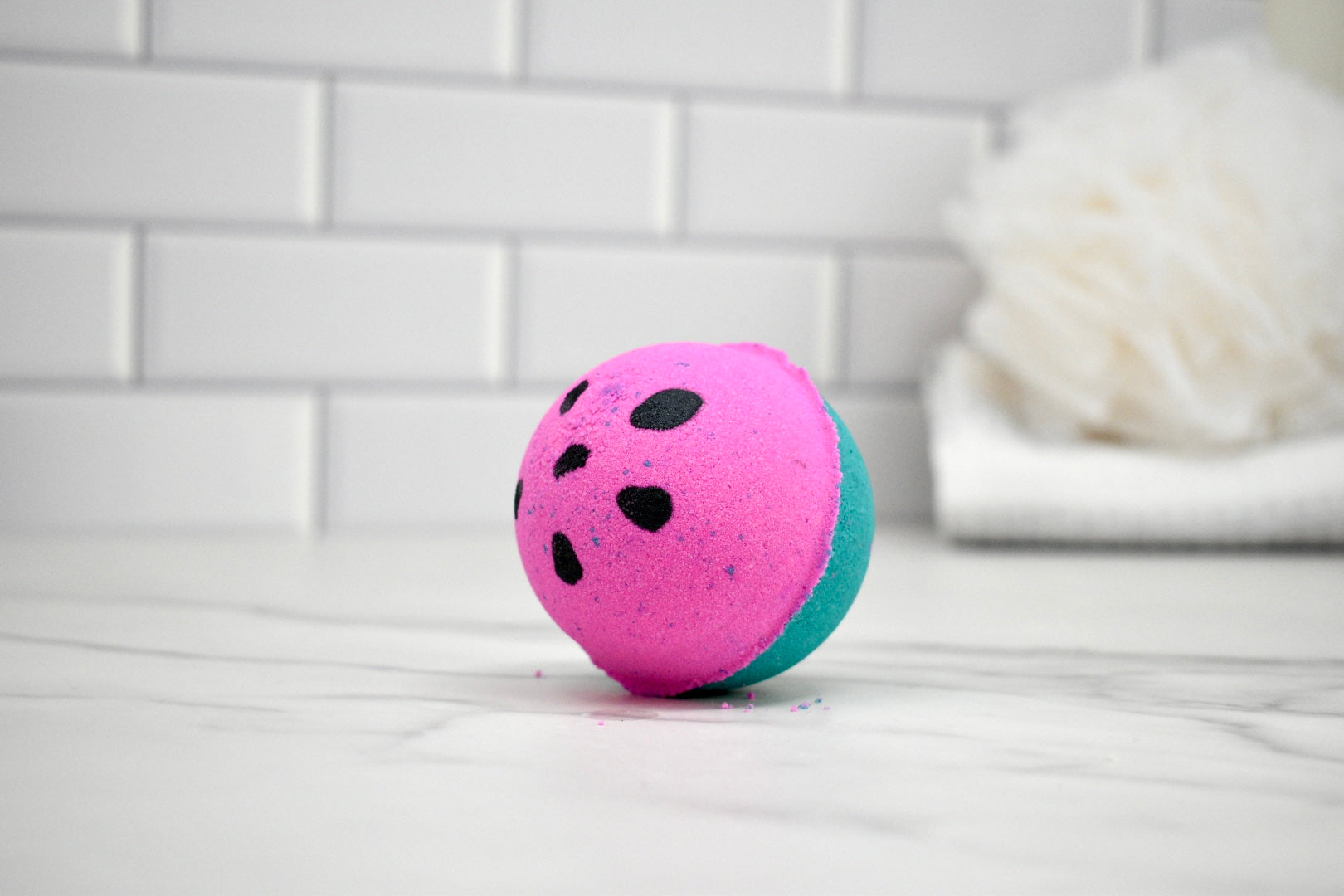 Pink and green bath bomb with black spots on a white tiled bathroom floor.