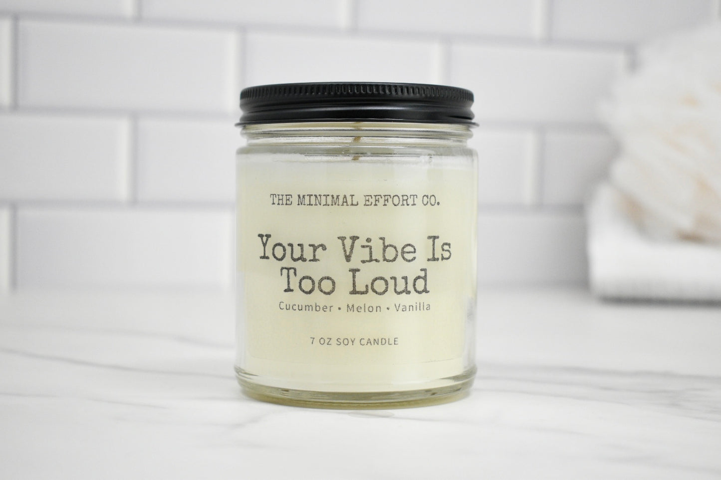 Candle jar labeled 'Your Vibe Is Too Loud' by The Minimal Effort Co. on a white surface with a blurred background.