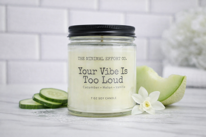 Candle jar labeled 'Your Vibe Is Too Loud' with cucumber slices and a flower on a white background